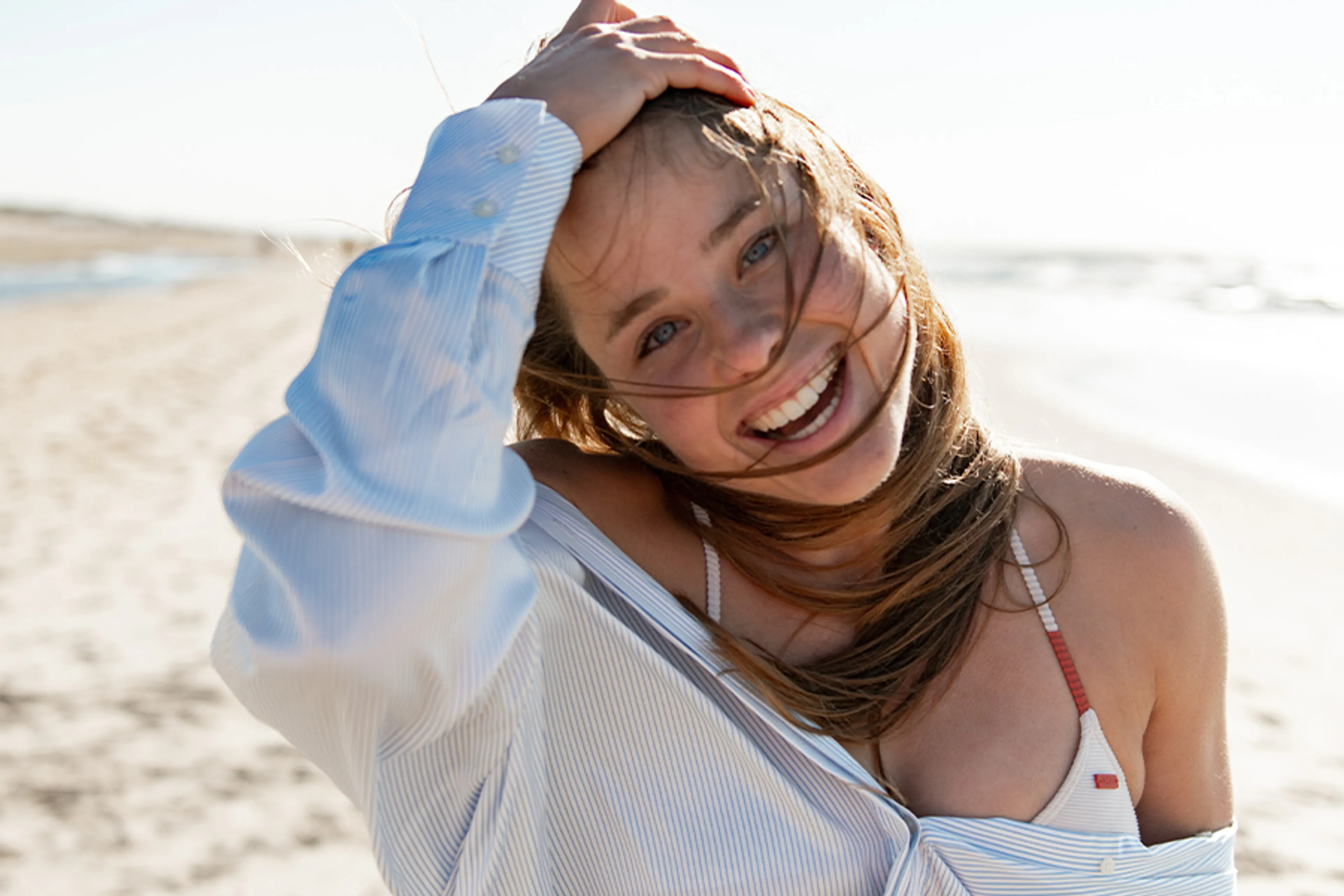 Een jonge vrouw die over het strand loopt in een bikini met een blouse daar over heen. Ze veegt met haar hand door haar haren en lacht naar de camera.